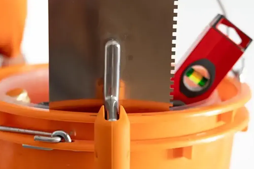 Orangefarbener Werkzeugeimer mit Fliesenkelle, Zahnspachtel und roter Wasserwaage auf hellem Hintergrund