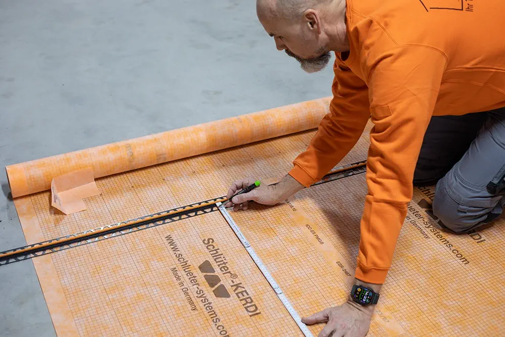 Mann in orangefarbenem Pullover misst Schlüter-KERDI Abdichtungsbahn mit Zollstock auf grauem Boden