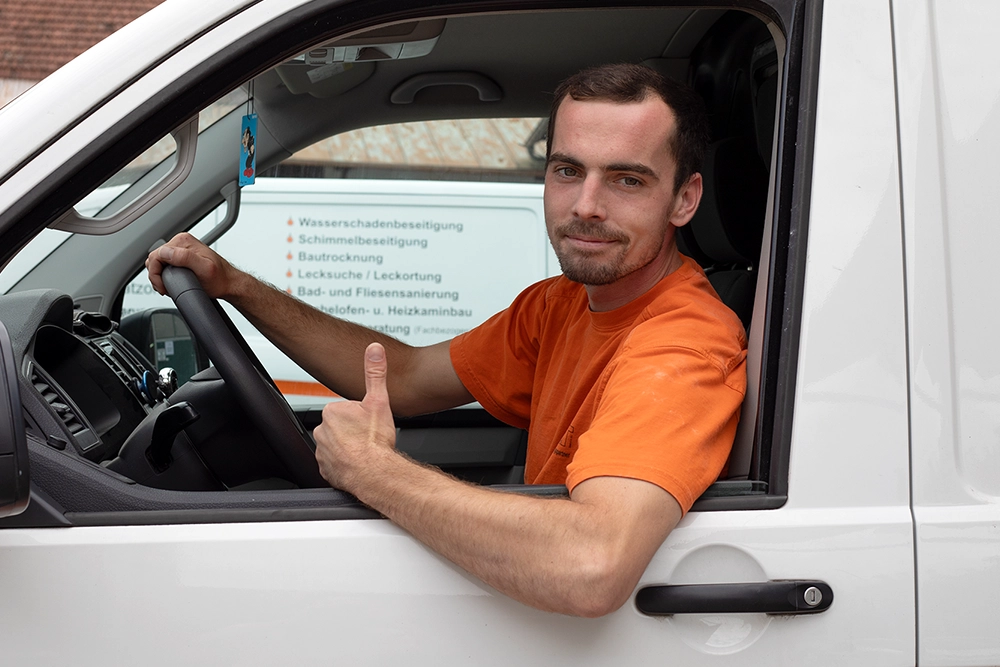 Junger Mann in orangefarbenem T-Shirt sitzt im weißen Firmenwagen und zeigt Daumen hoch