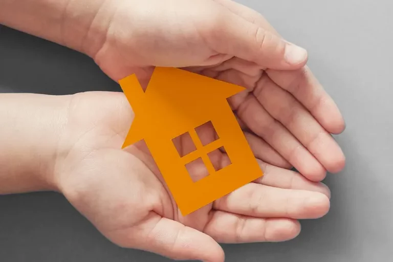 Kinderhände und Erwachsenenhände halten gemeinsam ein orangefarbenes Papierhaus vor grauem Hintergrund