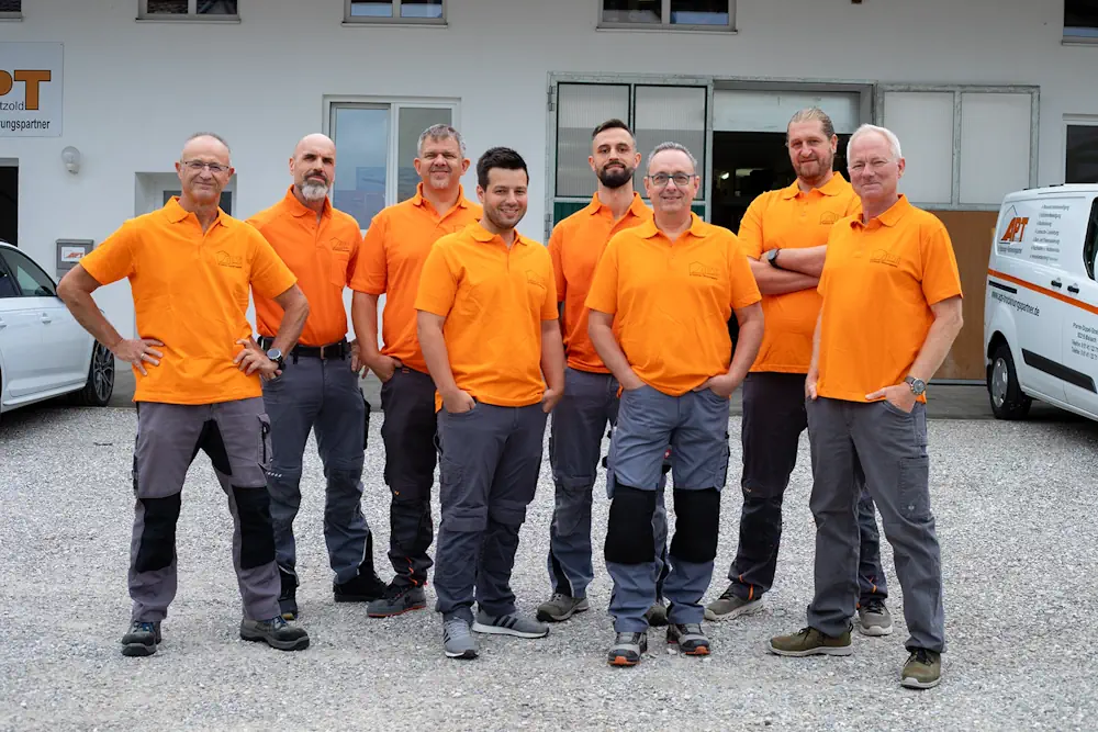 Acht Mitarbeiter in orangefarbenen Poloshirts und grauen Arbeitshosen stehen als Team vor einem Firmengebäude