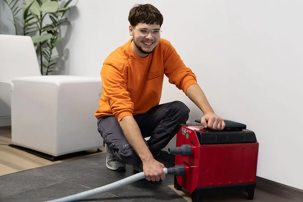Junger Techniker in orangem Firmen-Pullover kniet lächelnd auf dunklem Fliesenboden und bedient eine rote Rohrreinigungsmaschine