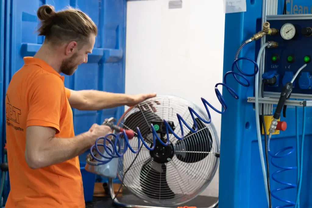 Techniker mit Dutt in orangem Poloshirt reinigt einen Standventilator mit Druckluft in blauer Werkstatt